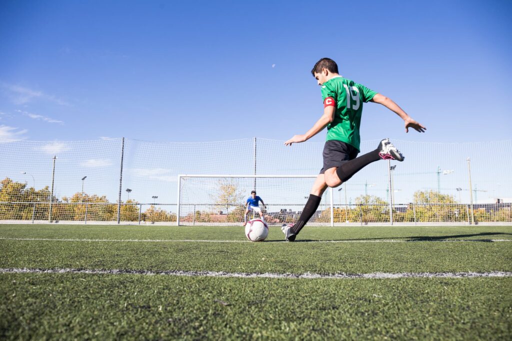 A man in the process of kicking a soccer ball towards a goalie in a net